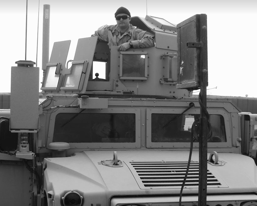 A black and white photo of a man wearing sunglasses and a beanie, leaning out of the turret of an armored Humvee. Various military equipment and sensors are mounted on the vehicle's exterior. A black and white photo of a man wearing sunglasses and a beanie, leaning out of the turret of an armored Humvee. Various military equipment and sensors are mounted on the vehicle's exterior.