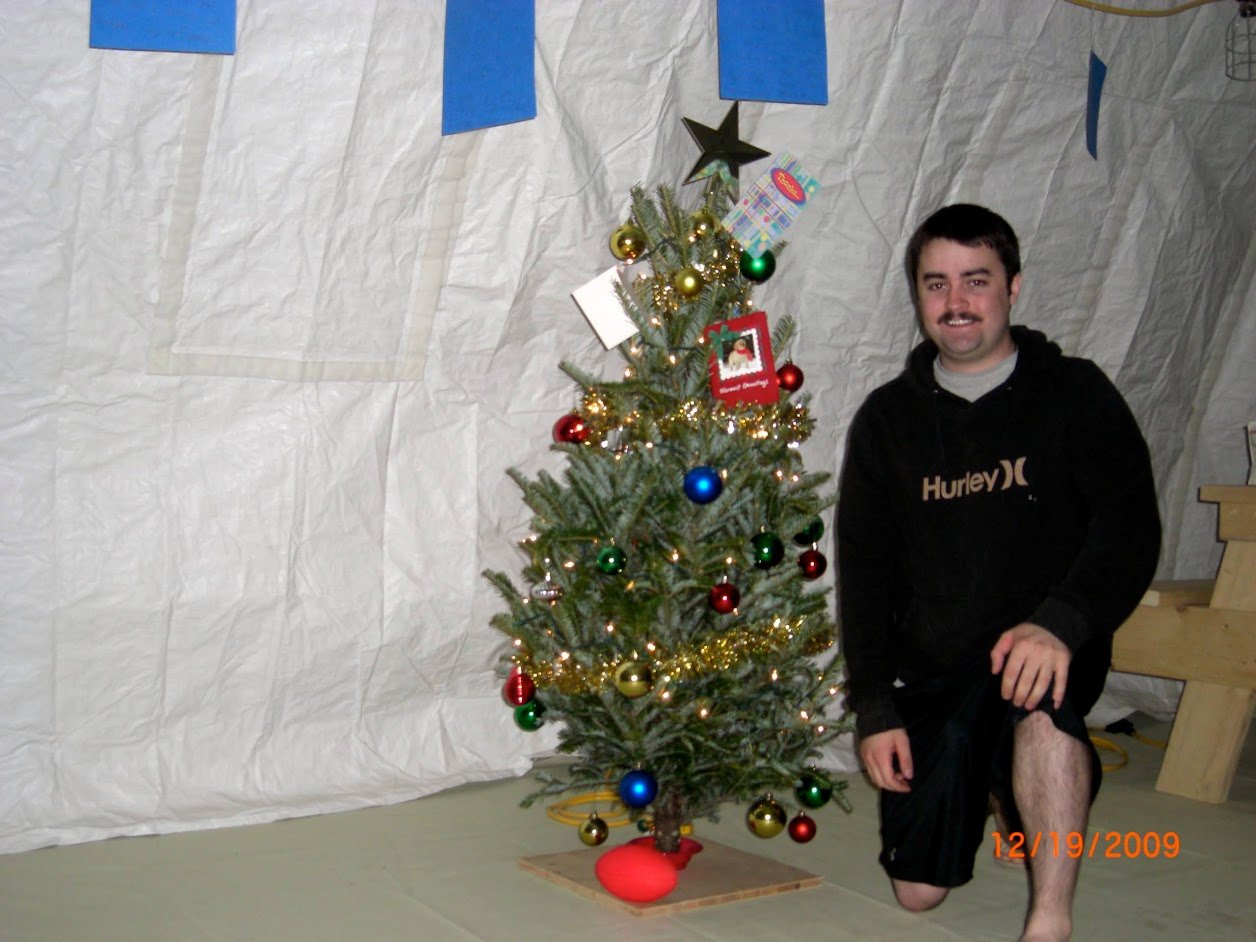 Tom kneeling next to a decorated Christmas tree inside the morale tent on December 19, 2009. The tree is adorned with gold tinsel, colorful ornaments, and holiday cards. A black star sits at the top. Tom is wearing a black Hurley hoodie. Tom kneeling next to a decorated Christmas tree inside the morale tent on December 19, 2009. The tree is adorned with gold tinsel, colorful ornaments, and holiday cards. A black star sits at the top. Tom is wearing a black Hurley hoodie.